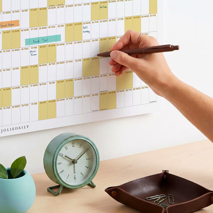 May include: A wall calendar with yellow highlighted weekends and handwritten notes, a brown ballpoint pen, a round mint green analog alarm clock, and a dark brown leather valet tray containing silver paper clips on a light wooden surface.