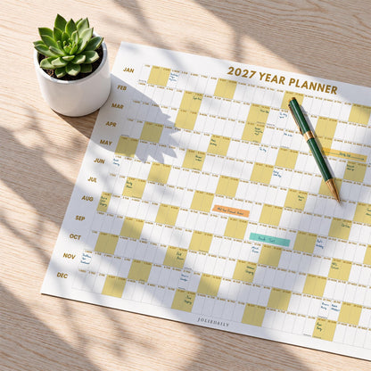May include: A 2027 year planner wall calendar with a grid layout featuring months from January to December, highlighted yellow columns, and handwritten notes. A green and gold pen rests on the paper, and a small green succulent in a white pot sits nearby.