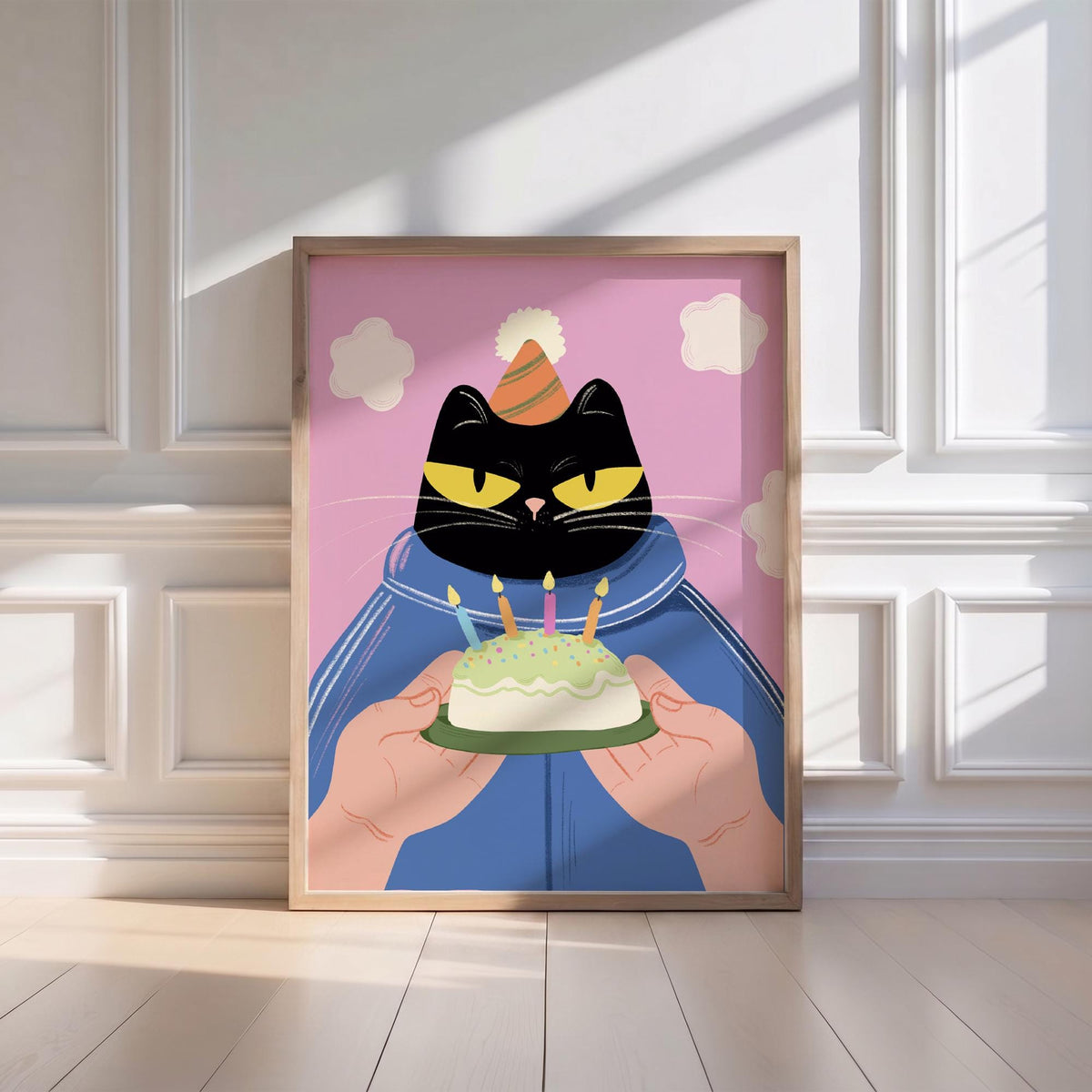 May include: A framed illustration features a black cat wearing a party hat and blue jacket, holding a small cake with lit candles. The background is pink with white cloud shapes. The artwork is in a wooden frame.
