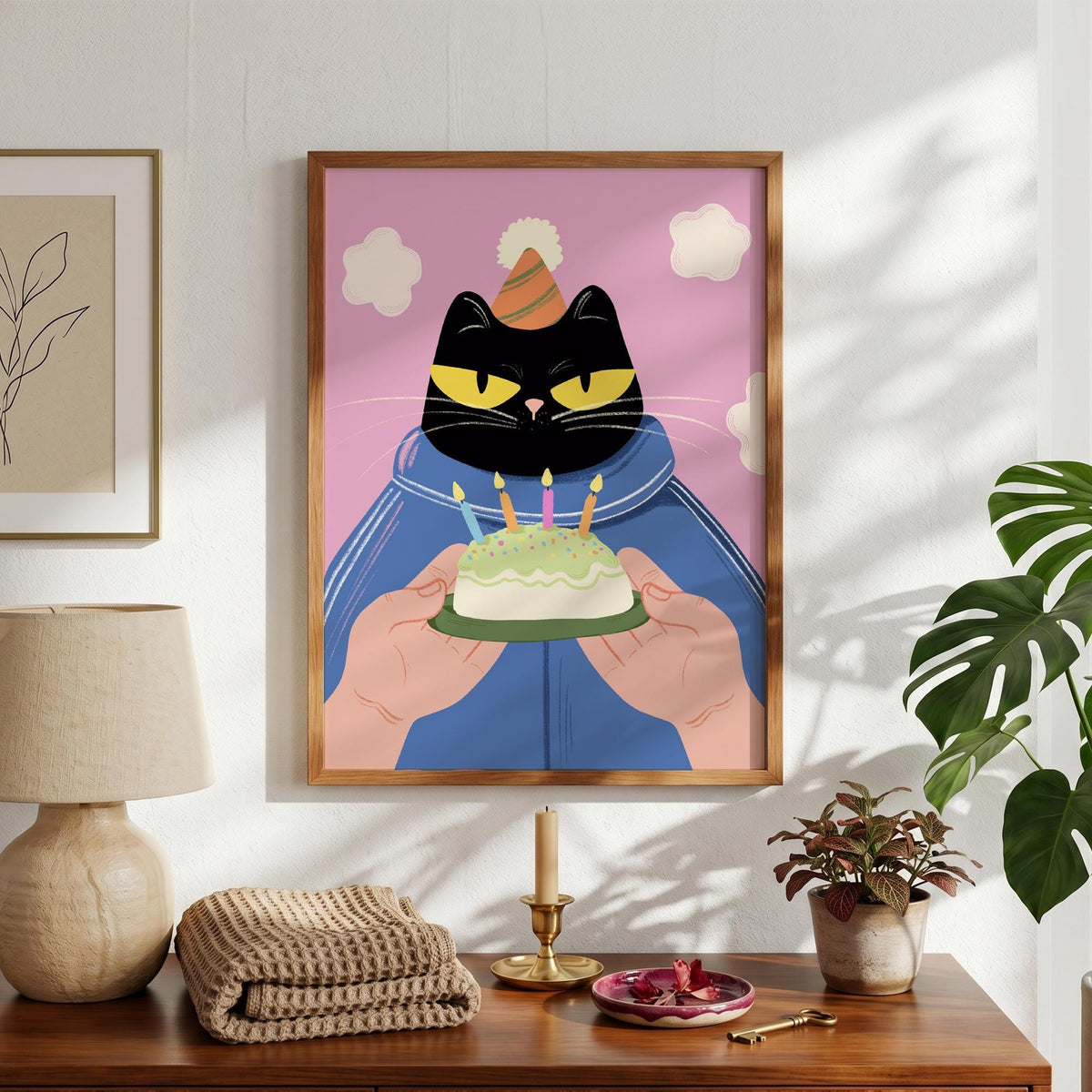 May include: A framed art print featuring a black cat wearing a party hat and a blue jacket. The cat holds a small cake with lit candles. The background is pink with white cloud shapes.
