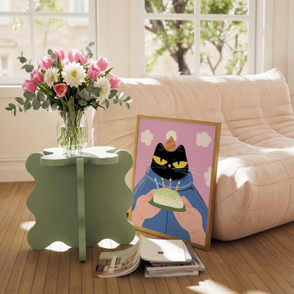 May include: A bouquet of pink tulips and white dahlias in a clear glass vase sits on a light green, four-leaf clover-shaped side table. A framed illustration of a black cat holding a birthday cake is propped against a sofa.