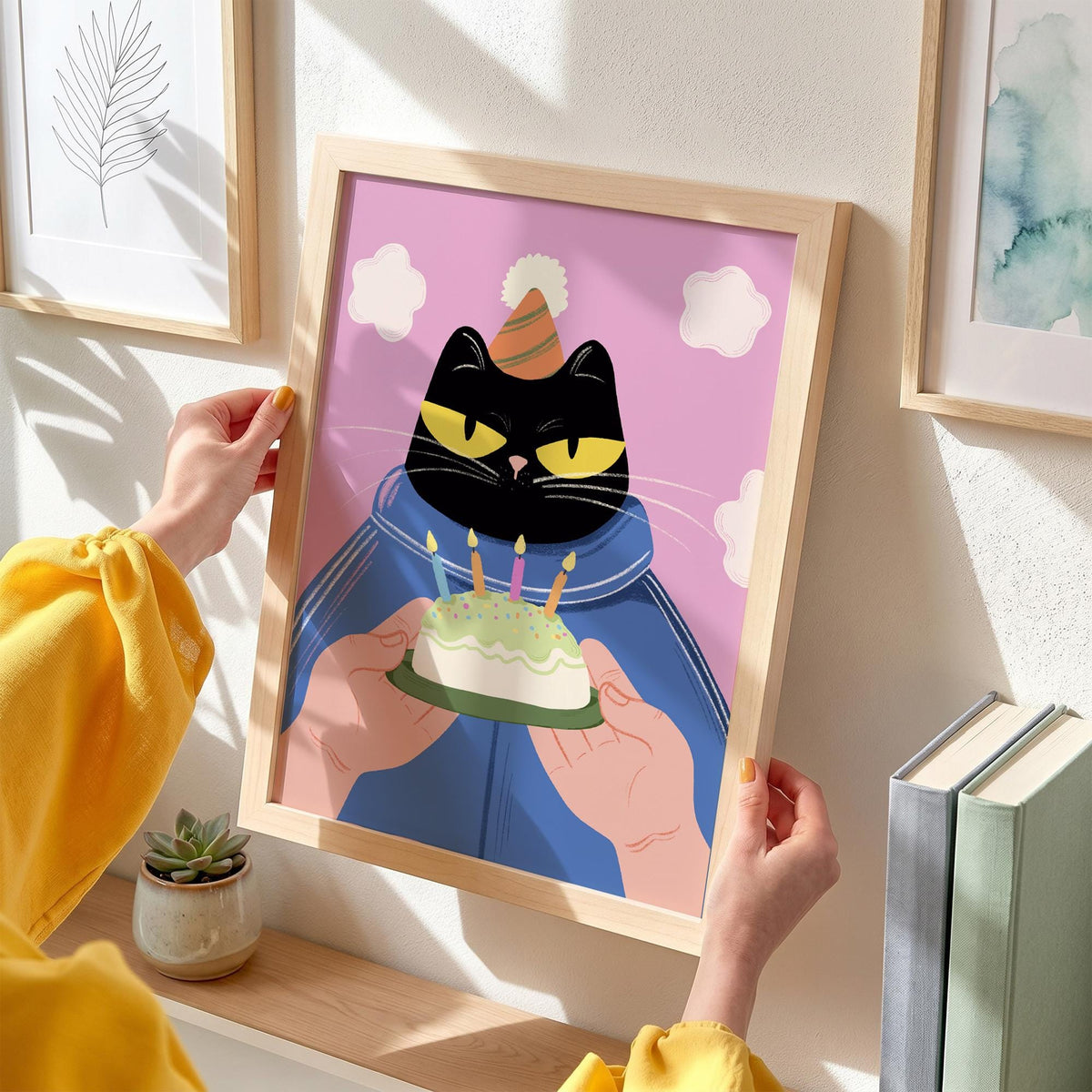 May include: A framed art print featuring a black cat wearing a party hat and blue jacket, holding a birthday cake with lit candles. The background is pink with white cloud shapes. The print is in a light wood frame.