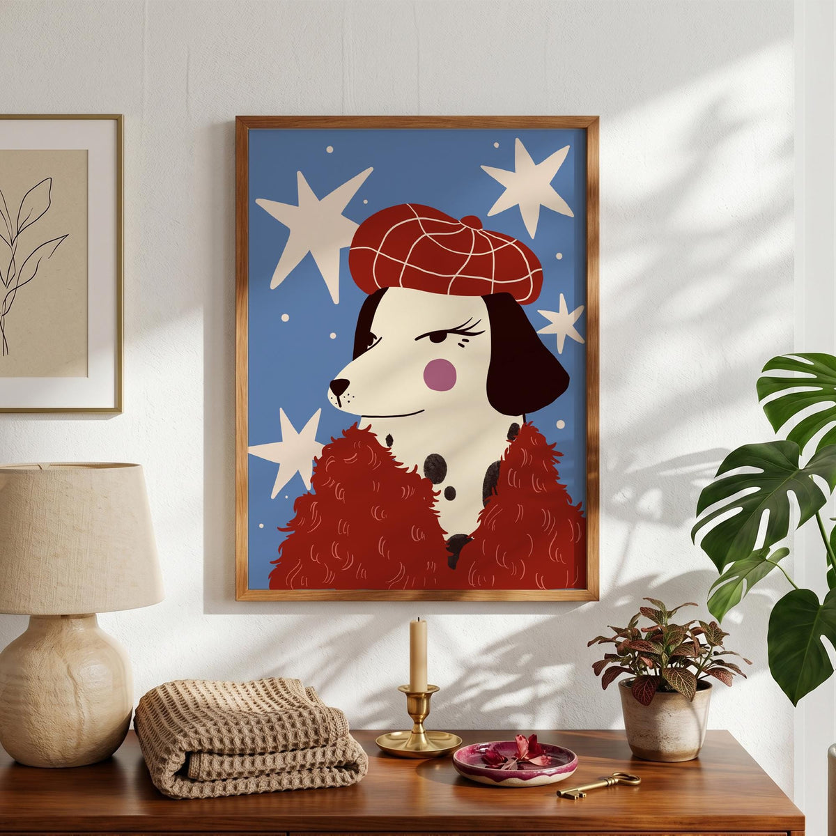 May include: A framed art print featuring a Dalmatian dog wearing a red beret and a red fluffy scarf against a blue background with white star shapes. The dog has black spots, a pink cheek, and a stern expression. The frame is a warm brown color.