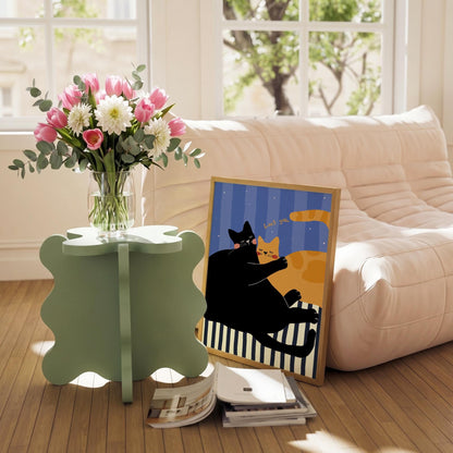 May include: A light green, four-leaf clover-shaped side table holds a clear glass vase filled with pink tulips, white dahlias, and eucalyptus. A framed art print depicts two cats with the text "Loves you." A stack of books rests on the floor.