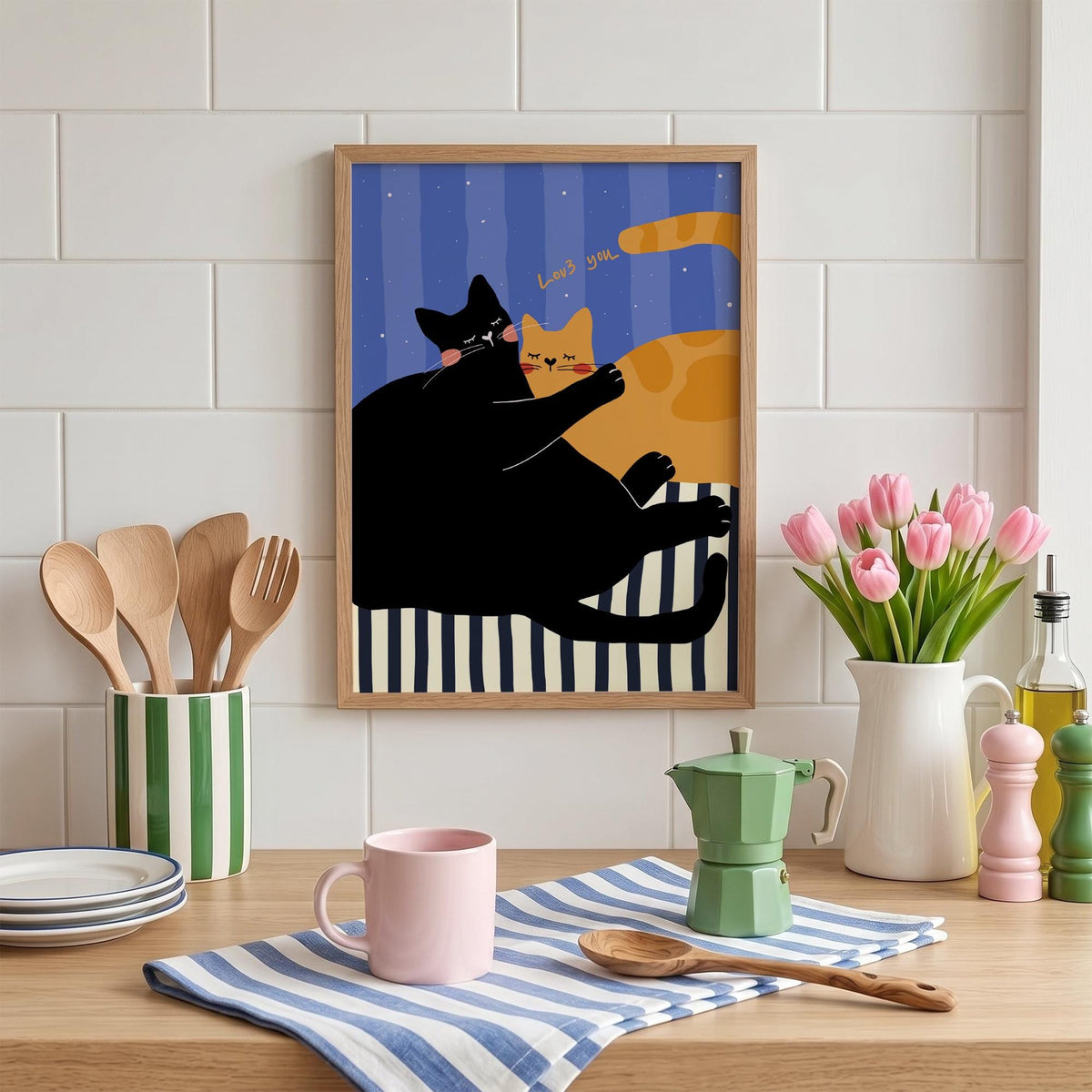 May include: A framed art print featuring two cartoon cats, one black and one orange, embracing. The background has blue vertical stripes and the words "Loves you". The print is in a wooden frame and is displayed on a wooden surface.