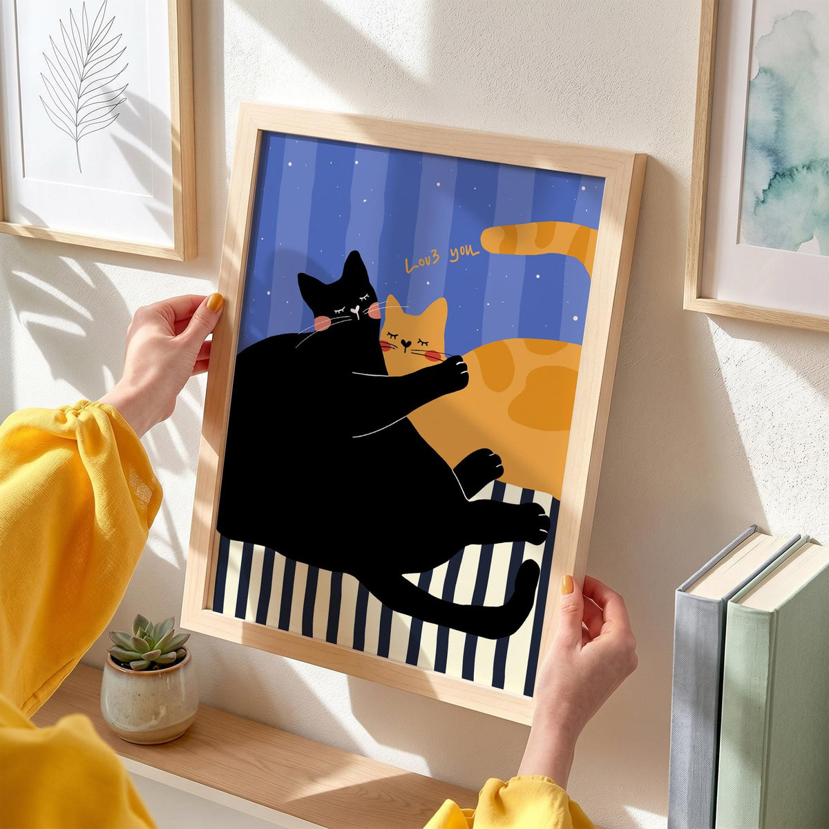 May include: A framed art print featuring two cats embracing. One cat is black, the other is orange, set against a blue striped background. The print includes the text "Loves you". The frame is made of light-colored wood.