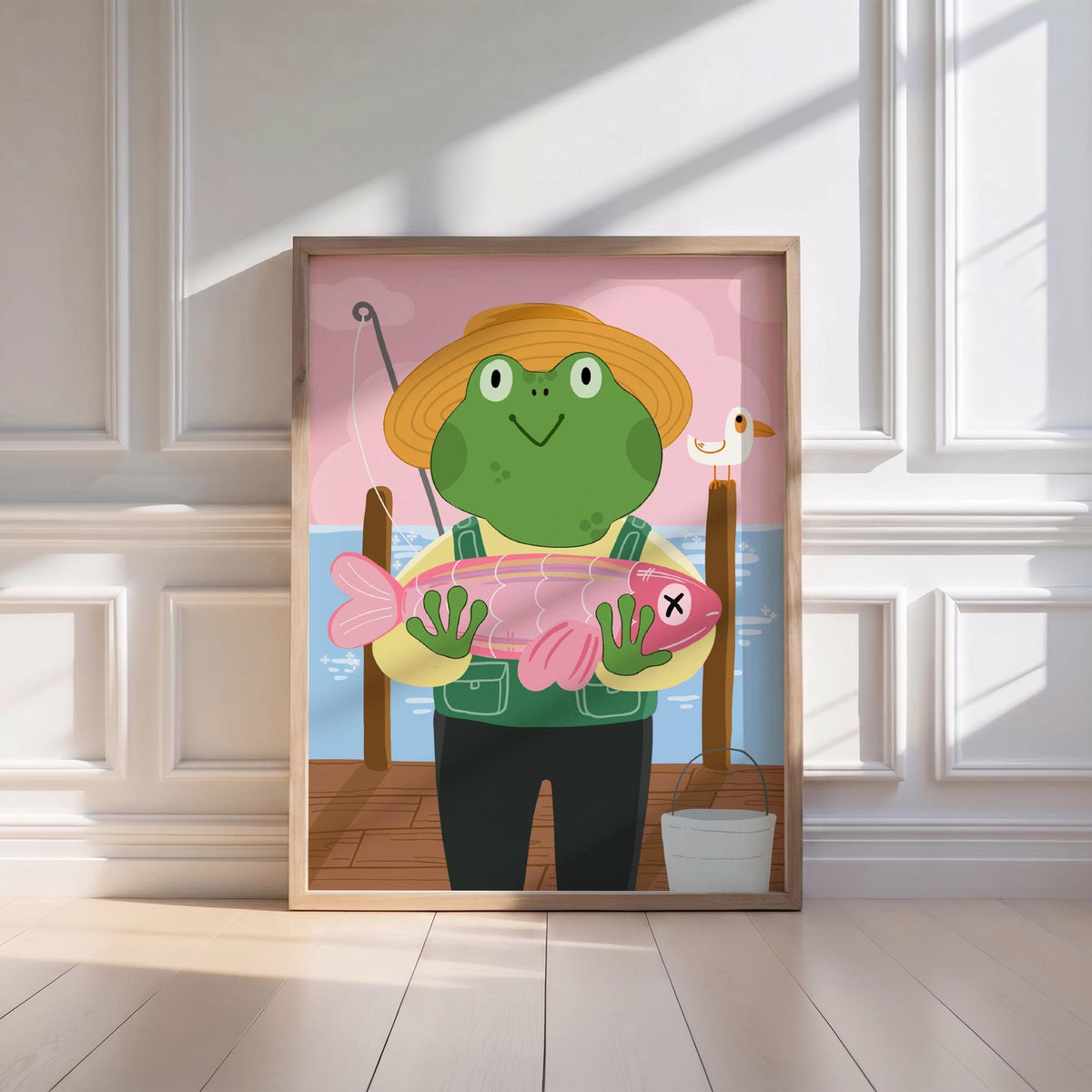 May include: A framed illustration of a cartoon frog wearing a straw hat and overalls, holding a pink fish with an X on it. The frog stands on a wooden dock with a fishing rod, a seagull, and a bucket. The background features a pink sky and blue water.