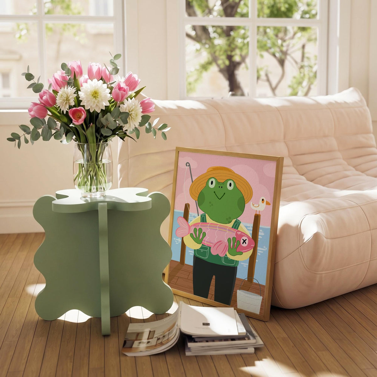 May include: A light green, four-leaf clover-shaped side table holds a clear glass vase filled with pink tulips, white dahlias, and eucalyptus. A framed illustration of a frog fishing is propped against a beige sofa.