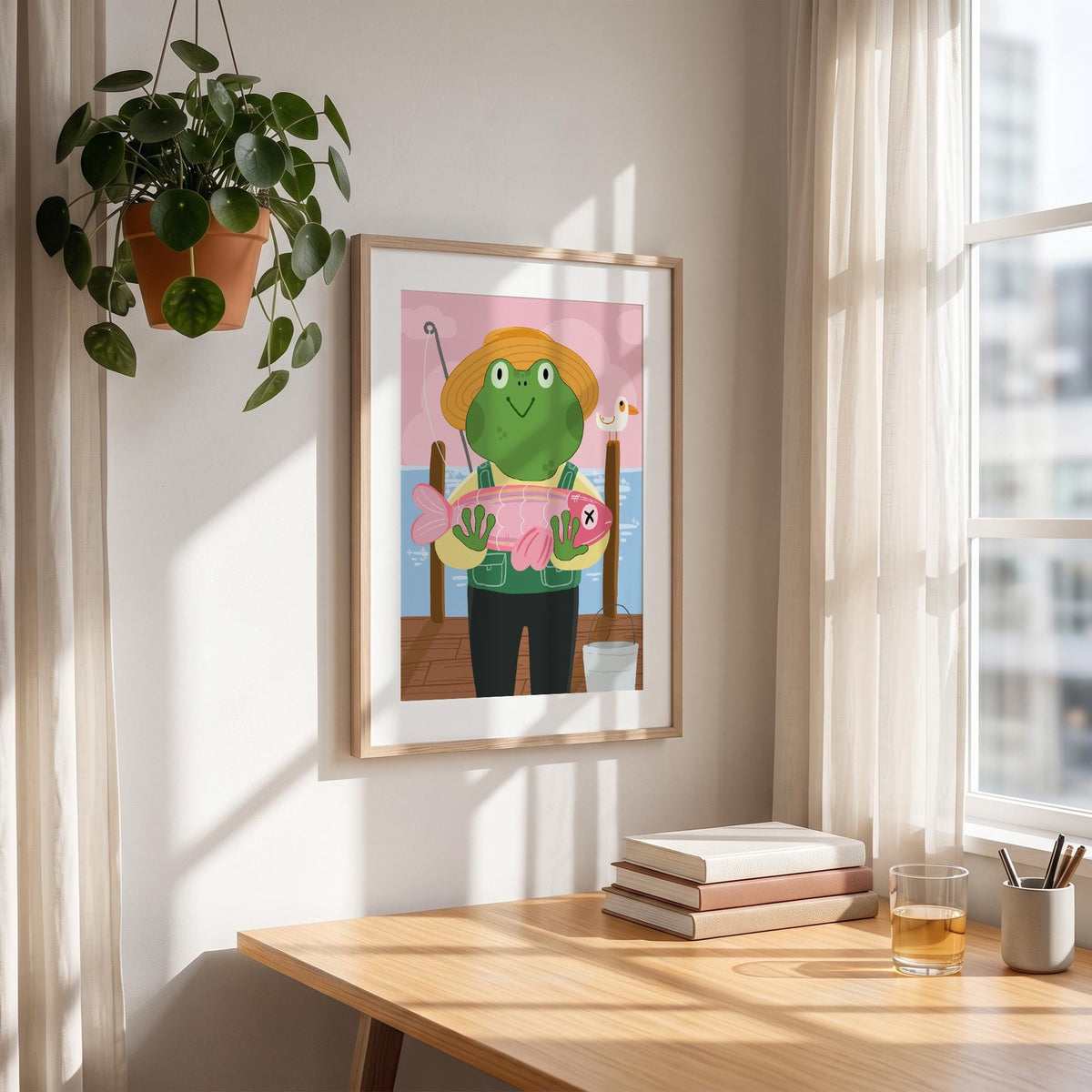 May include: A framed art print featuring a cartoon frog wearing a straw hat and vest, holding a pink fish with an X on it. The background includes a pier, water, and a pink and blue sky. A hanging plant and books are in the foreground.