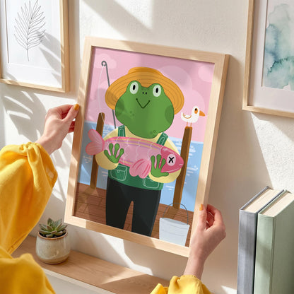 May include: A framed print featuring a cartoon frog wearing a straw hat and overalls, holding a pink fish with an X mark. The background shows a pier, water, and a small bird. The frame is light wood.