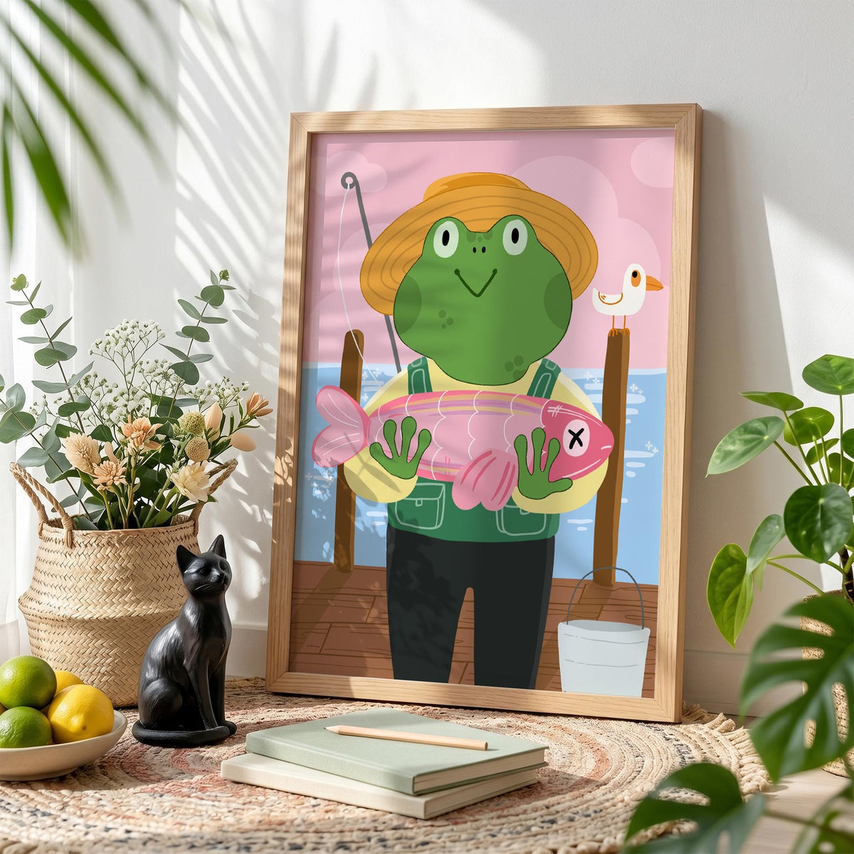 May include: A framed illustration of a cartoon frog wearing a straw hat and overalls, holding a pink fish with an X on it. The background shows a pier, water, and a pink sky. A small white bucket is visible.