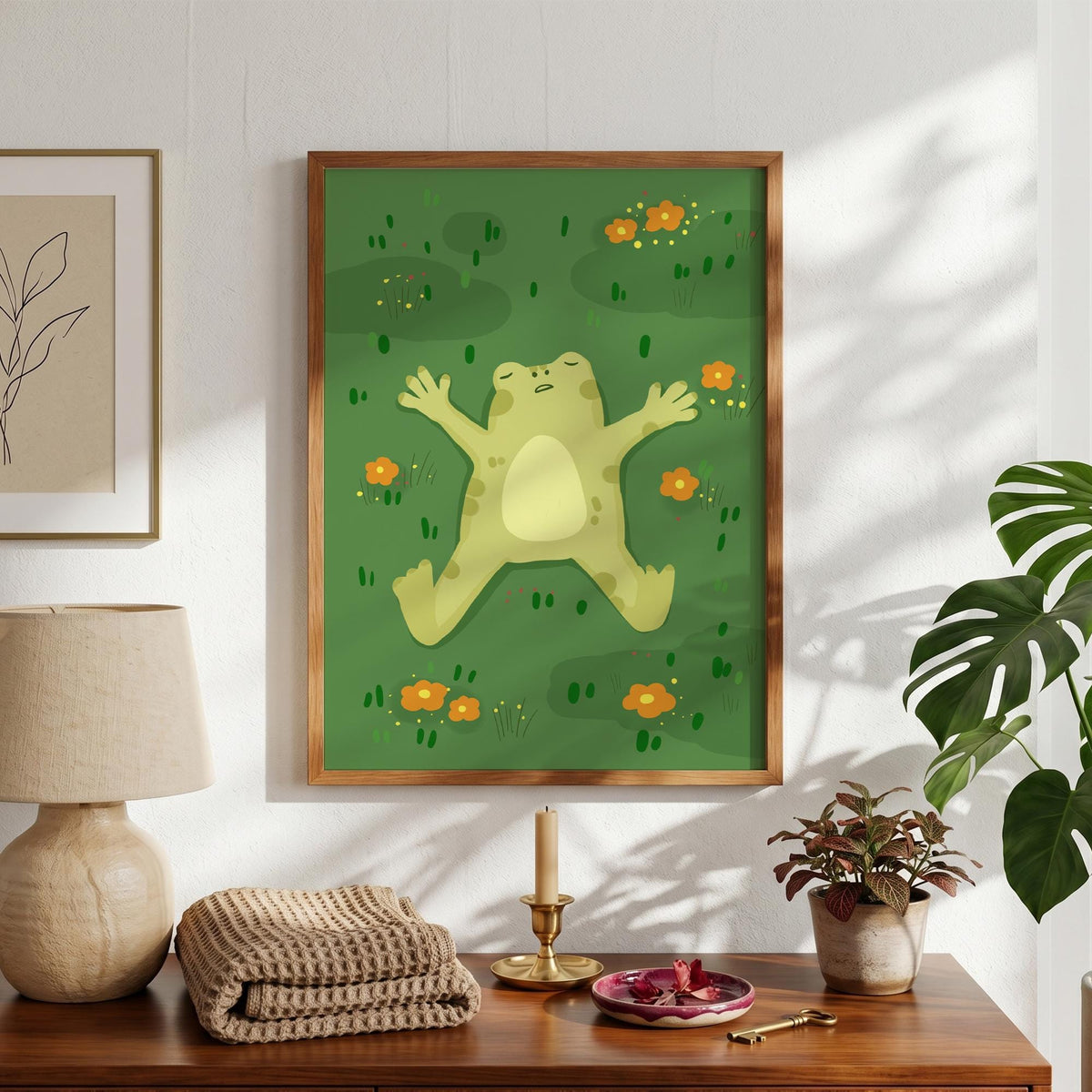 May include: A framed art print featuring a light green frog lying on its back in a field of green grass with orange flowers. The frog has its arms and legs spread out. The artwork is in a wooden frame.