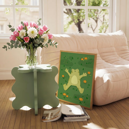 May include: A light green, four-leaf clover-shaped side table holds a clear glass vase filled with pink tulips, white dahlias, and eucalyptus. A framed print of a cartoon frog on a green background rests nearby. A stack of magazines sits on the floor.