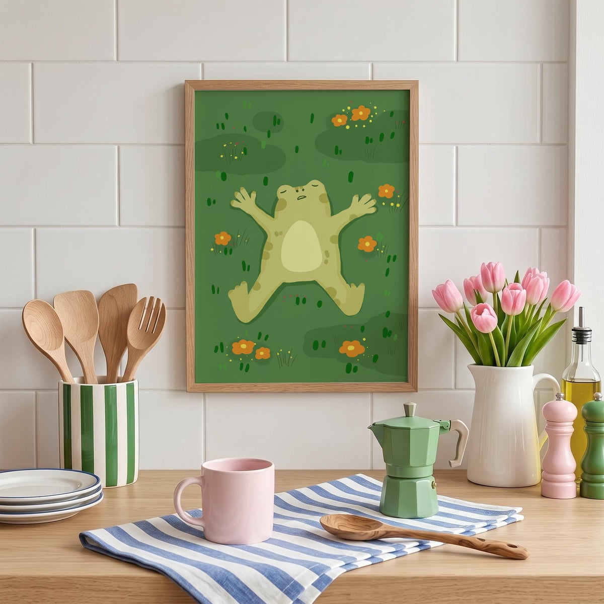 May include: A framed art print featuring a cartoon frog relaxing on a green background with orange flowers. The frog is light green with a cream-colored belly. The frame is a light brown color. The print is displayed on a wooden surface.