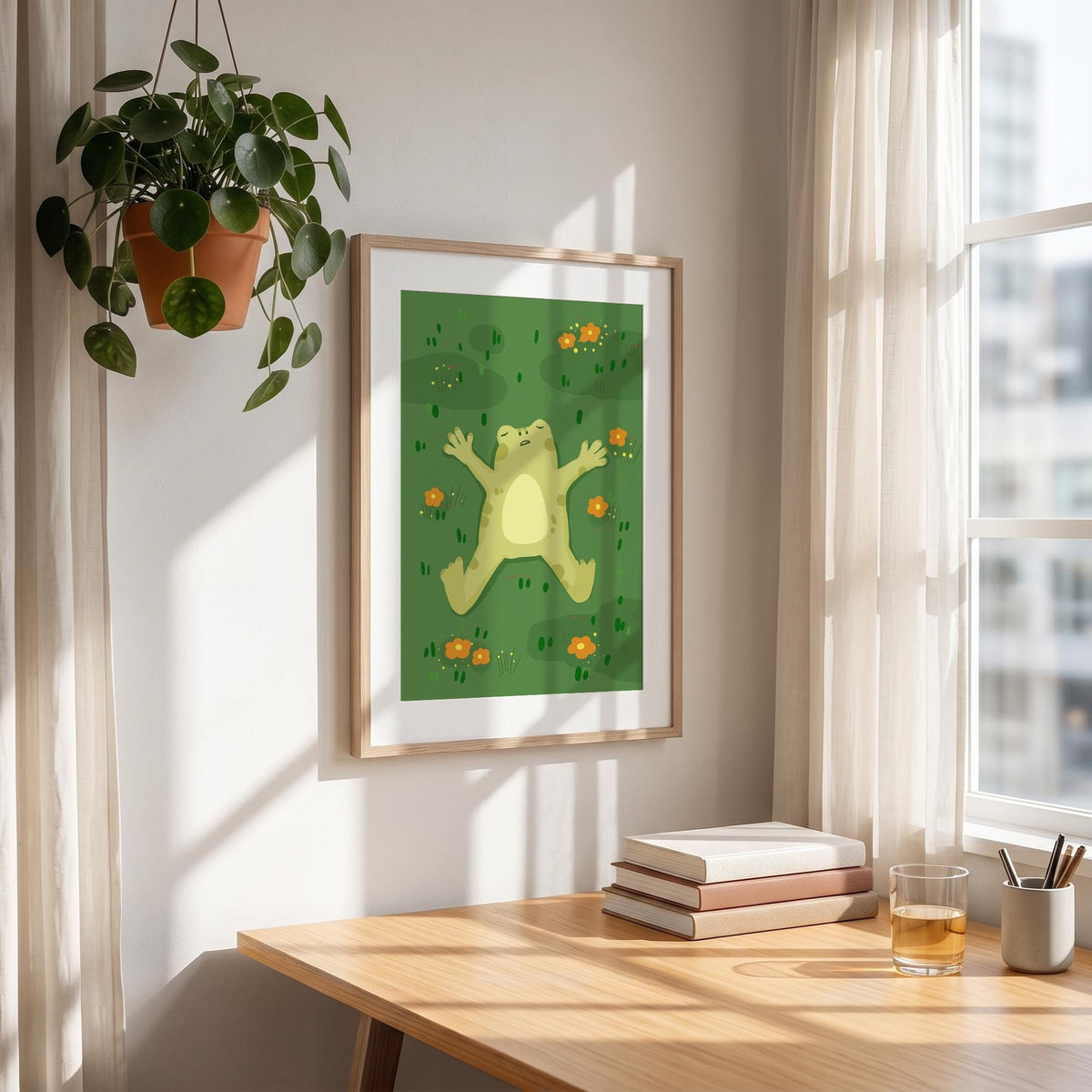 May include: A framed art print featuring a cartoon frog lying on its back in a field of green grass and orange flowers. The frog is light green with a yellow belly. The artwork is displayed on a wooden desk with books and a glass of liquid.