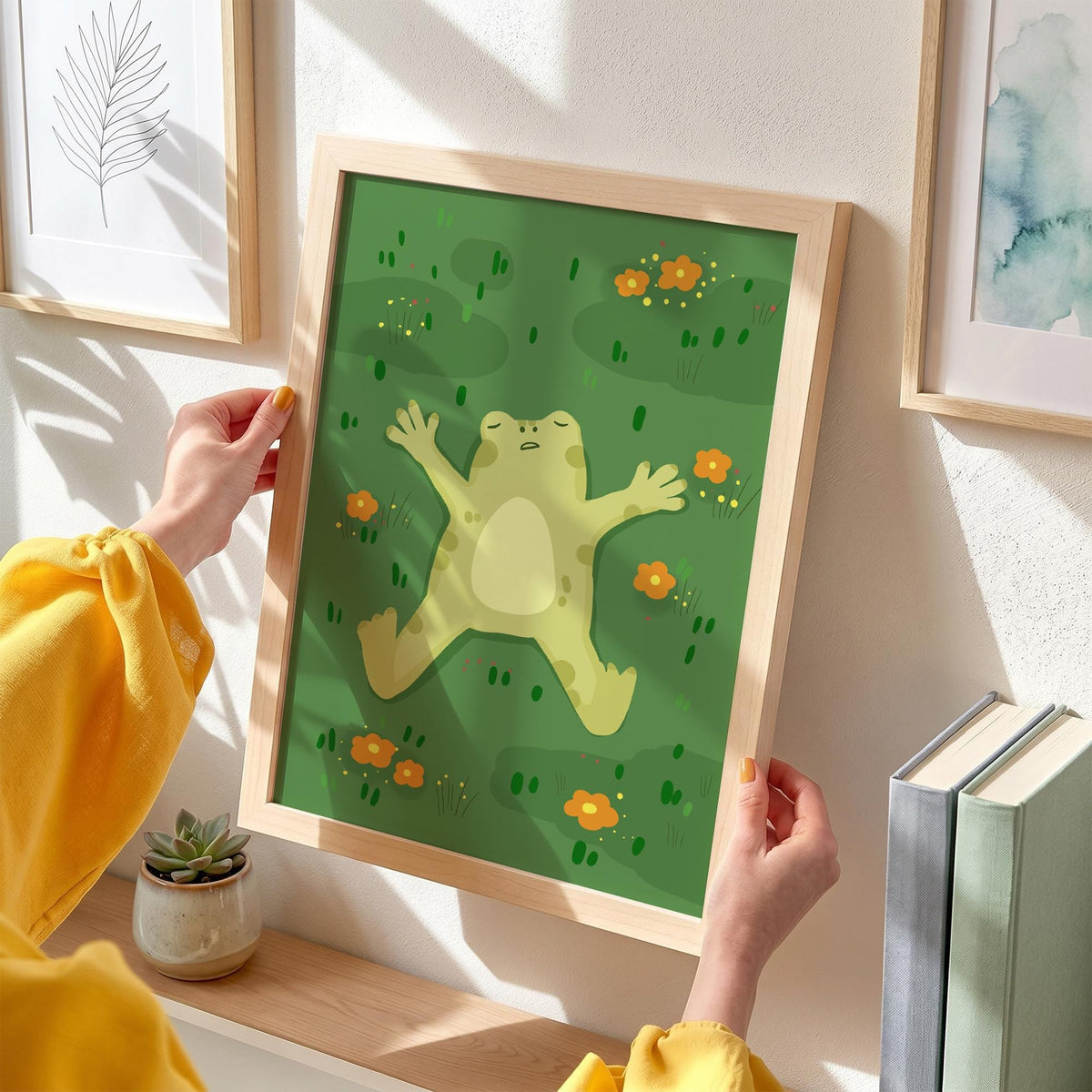 May include: A framed art print featuring a cartoon frog relaxing on a green background with orange flowers. The frog is light green with a cream-colored belly. The frame is made of light-colored wood. The art print is held by a person.