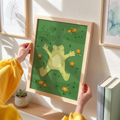 May include: A framed art print featuring a cartoon frog relaxing on a green background with orange flowers. The frog is light green with a cream-colored belly. The frame is made of light-colored wood. The art print is held by a person.