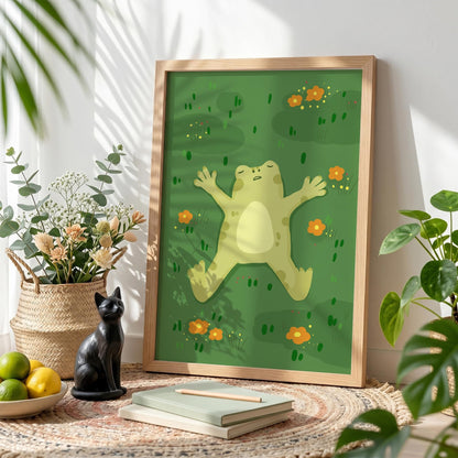 May include: A framed art print featuring a cartoon frog lying on its back in a green field with orange flowers. The artwork is in a light wooden frame, and the scene is set on a woven rug with a black cat statue and a bowl of citrus fruits.