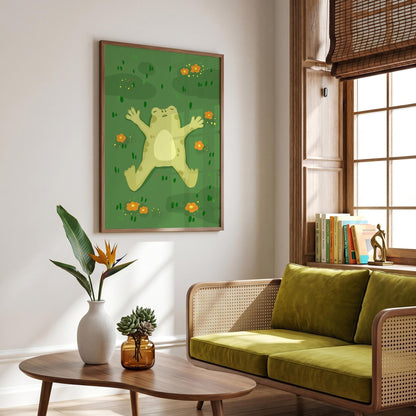 May include: A framed art print featuring a whimsical illustration of a light green frog lying on its back in a grassy field. The frog has its arms and legs outstretched. Small orange flowers are scattered around the frog. The artwork is framed in a brown wooden frame.