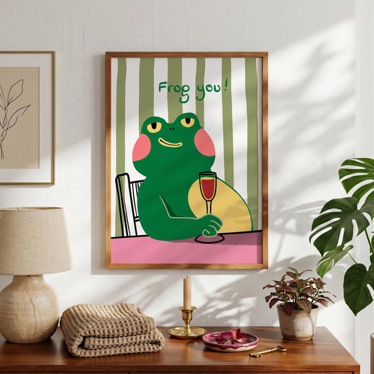 May include: A framed art print featuring a cartoon frog holding a glass of red wine. The frog is green with pink cheeks and yellow eyes, set against a striped green and white background. The text "Frog you!" is above the frog.