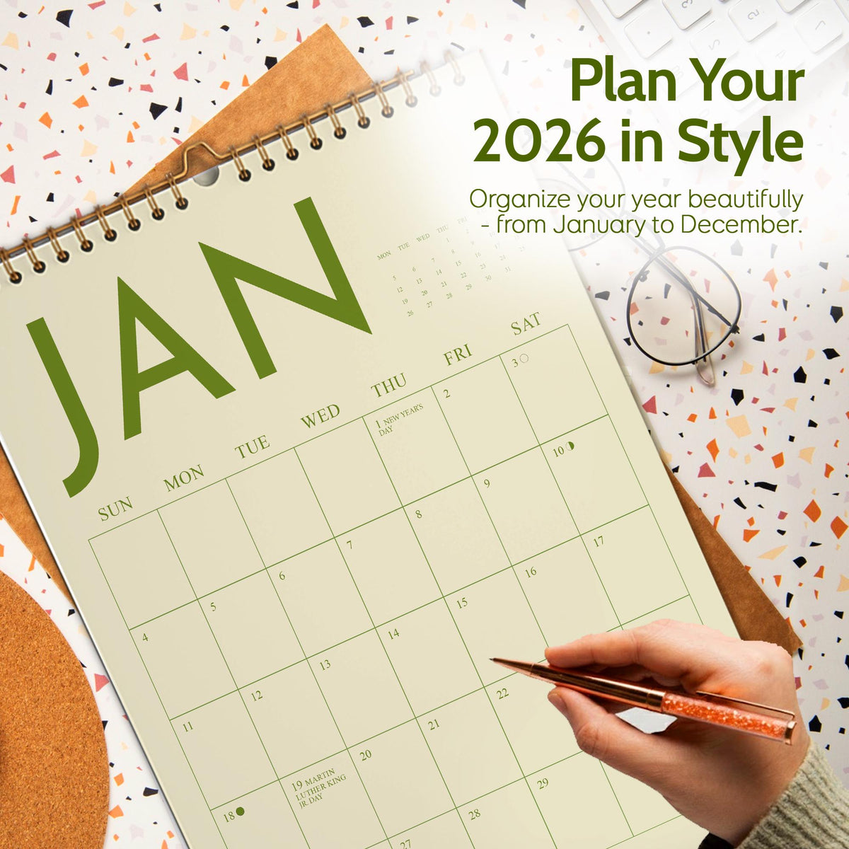 May include: A large, open calendar for the year 2026, with the month of January prominently displayed. The calendar is cream-colored with green lettering and a spiral binding. The text "Plan Your 2026 in Style" is at the top. A hand holds a pen.