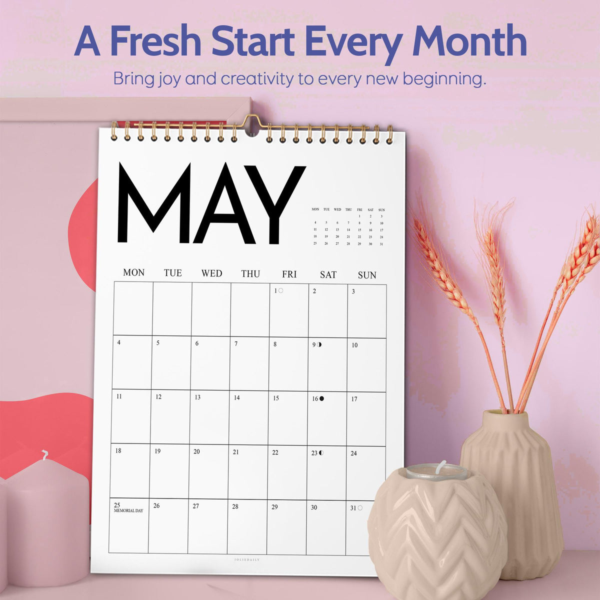 May include: A spiral-bound calendar open to May, with a gold-colored metal hook. The calendar features a simple grid layout with the days of the week across the top. The text "A Fresh Start Every Month" is at the top. Decorative vases and candles are in the foreground.