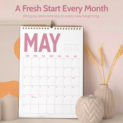May include: A white wall calendar for May with a gold spiral binding. The word "MAY" is in large pink letters. The calendar is on a white surface with a candle holder and vase with wheat stalks.