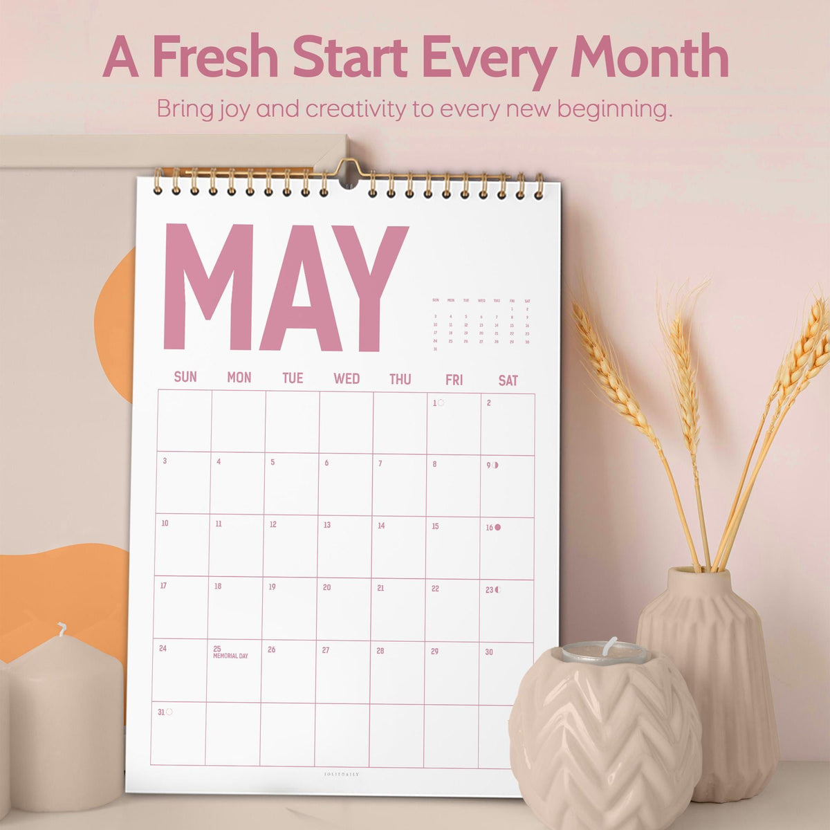 May include: A white wall calendar for May with a gold spiral binding. The word "MAY" is in large pink letters. The calendar is on a white surface with a candle holder and vase with wheat stalks.