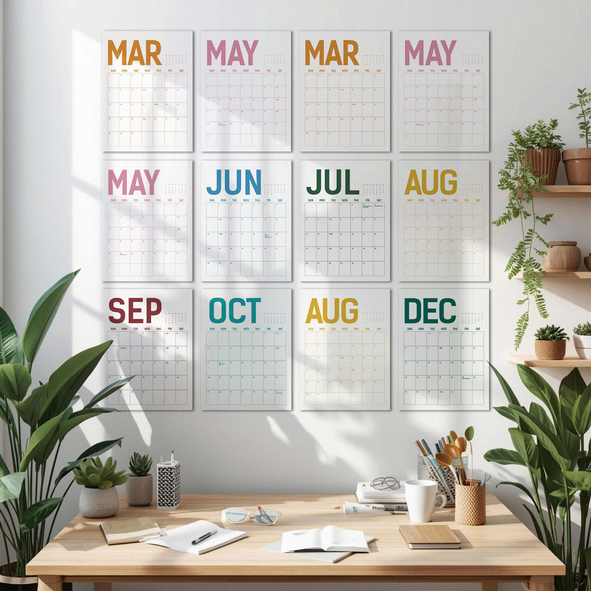 May include: A set of twelve monthly wall calendars, each with a different month's name in a vibrant color. The calendars are arranged in a grid on a white wall, with a wooden desk and plants in the foreground.