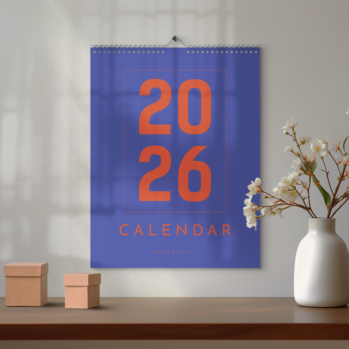 May include: A wall calendar for the year 2026, featuring a vibrant blue background with large orange numerals and the word "CALENDAR." The calendar is suspended by a silver spiral binding. Two small, brown cardboard boxes and a white vase with flowers are on a wooden surface.