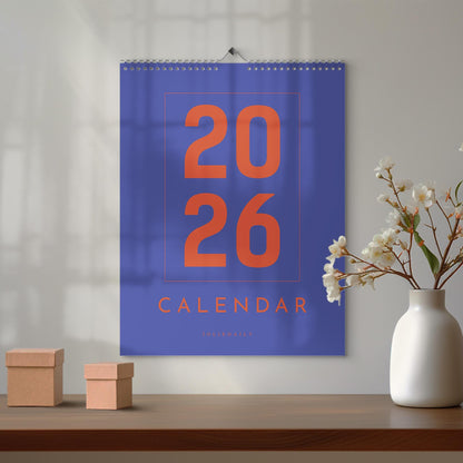 May include: A wall calendar for the year 2026, featuring a vibrant blue background with large orange numerals and the word "CALENDAR." The calendar is suspended by a silver spiral binding. Two small, brown cardboard boxes and a white vase with flowers are on a wooden surface.