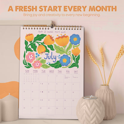 May include: A wall calendar for July with a floral design in orange, blue, and pink. The calendar has a gold spiral binding and includes the text "A FRESH START EVERY MONTH" and "Bring joy and creativity to every new beginning."