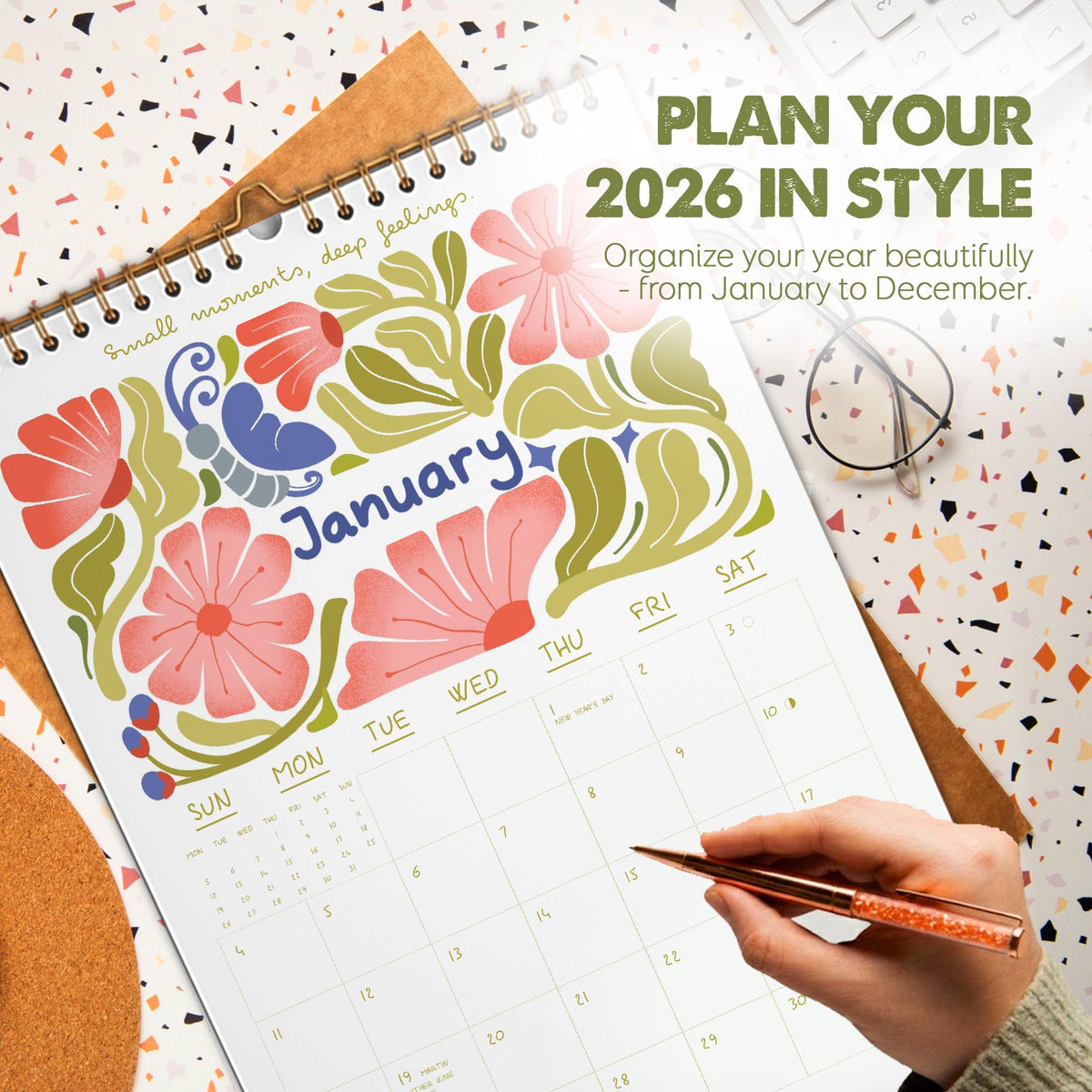 May include: A 2026 calendar open to January, featuring floral illustrations in pink, red, and green. The calendar has a spiral binding and includes the text "Plan Your 2026 in Style". A hand holds a pen, pointing to a date.