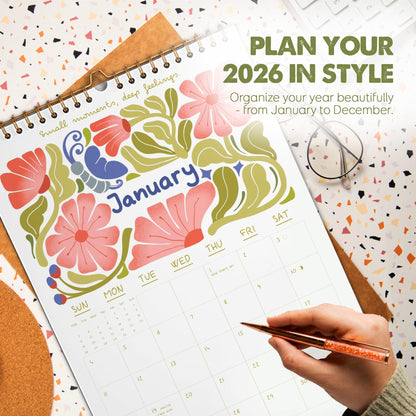 May include: A 2026 calendar open to January, featuring floral illustrations in pink, red, and green. The calendar has a spiral binding and includes the text "Plan Your 2026 in Style". A hand holds a pen, pointing to a date.