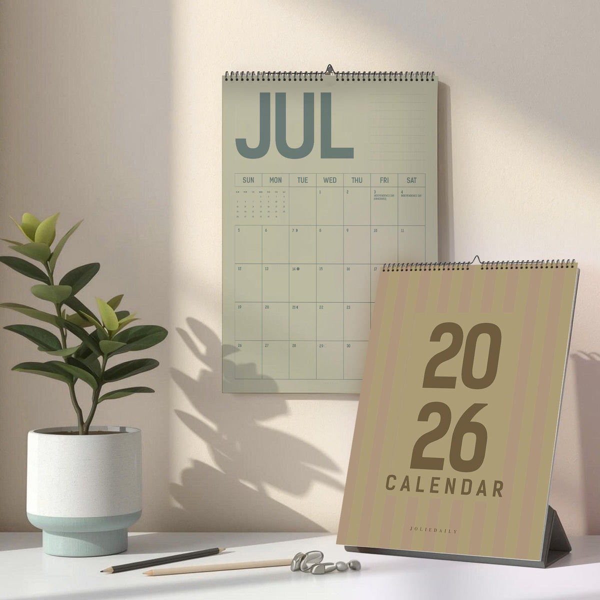 May include: A wall calendar displaying the month of July, and a desk calendar for the year 2026. The calendars are in shades of green and tan. A potted plant, pencils, and decorative stones are on the desk.