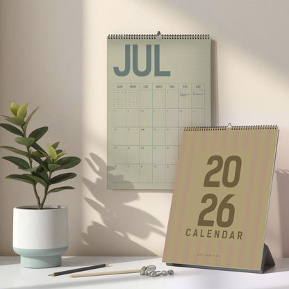May include: A wall calendar displaying the month of July, and a desk calendar for the year 2026. The calendars are in shades of green and tan. A potted plant, pencils, and decorative stones are on the desk.