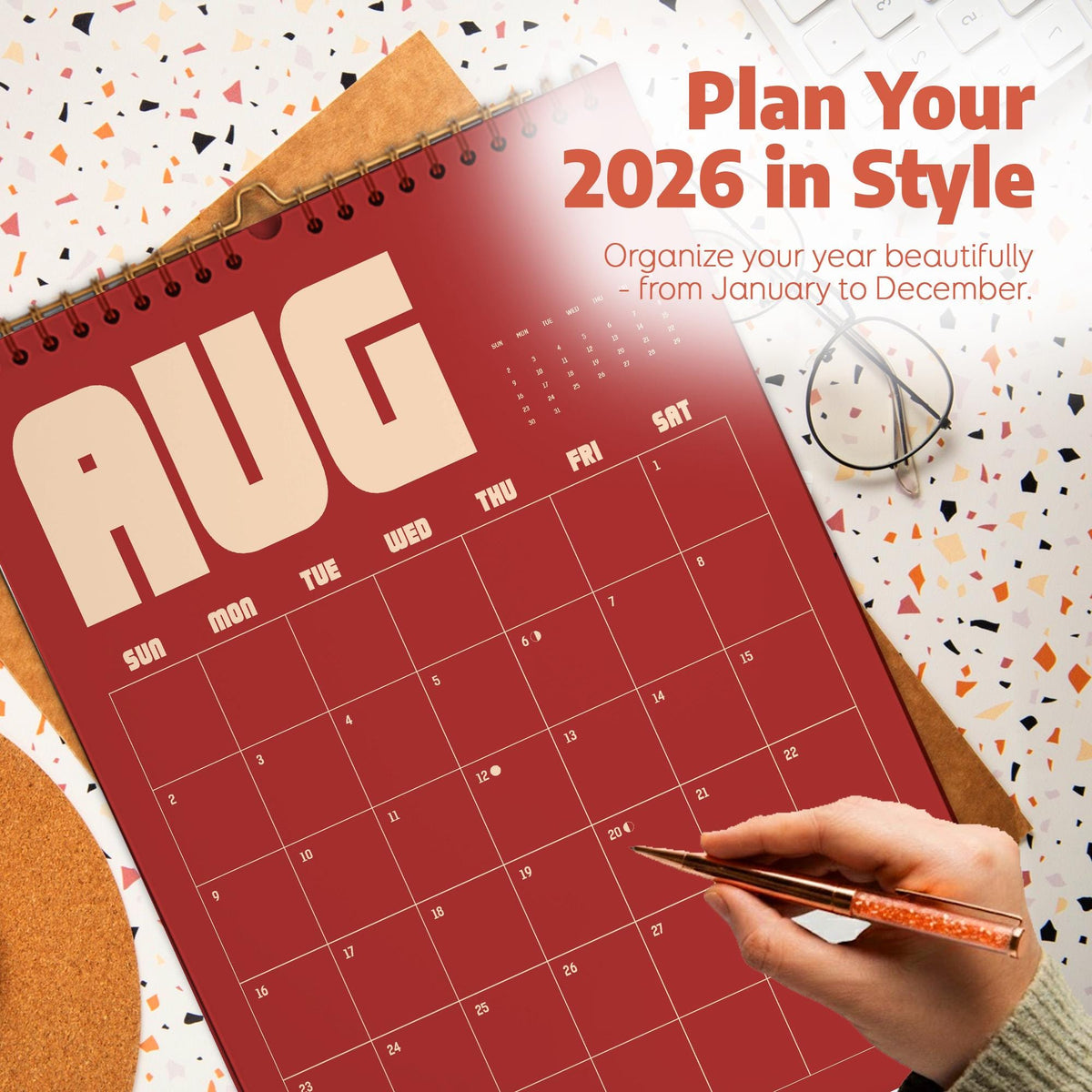 May include: A red 2026 calendar with the month of August displayed. The calendar is open, showing the days of the month. The text "Plan Your 2026 in Style" is at the top. A hand is holding a pen, ready to write on the calendar.