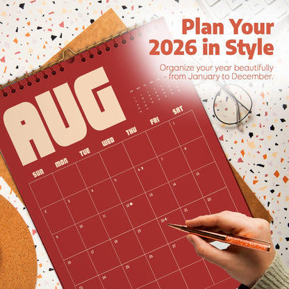 May include: A red 2026 wall calendar with the month of August displayed. The calendar has a spiral binding and is open to the month of August. The text "Plan Your 2026 in Style" is at the top. A hand is holding a pen.