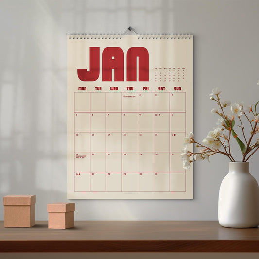 May include: A wall calendar for January with a cream background and red text. The word "JAN" is at the top, with the days of the week below. The calendar is hanging on a white wall, with a vase of flowers and two small boxes on a wooden surface.