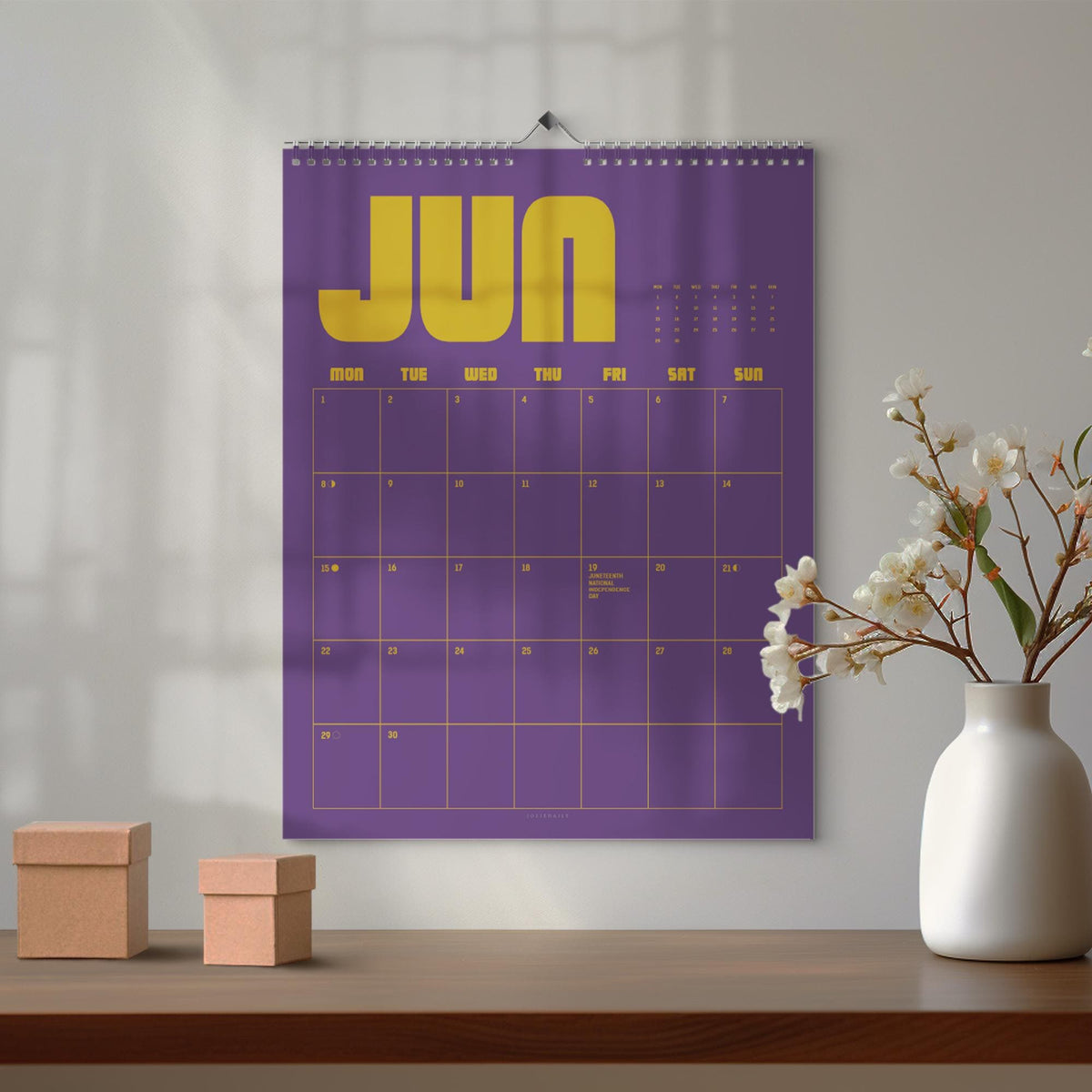 May include: A purple wall calendar for June with gold lettering and grid lines. The word "JUN" is prominently displayed at the top. The calendar is hanging on a white wall, with a white vase of flowers and two small brown boxes on a wooden surface.