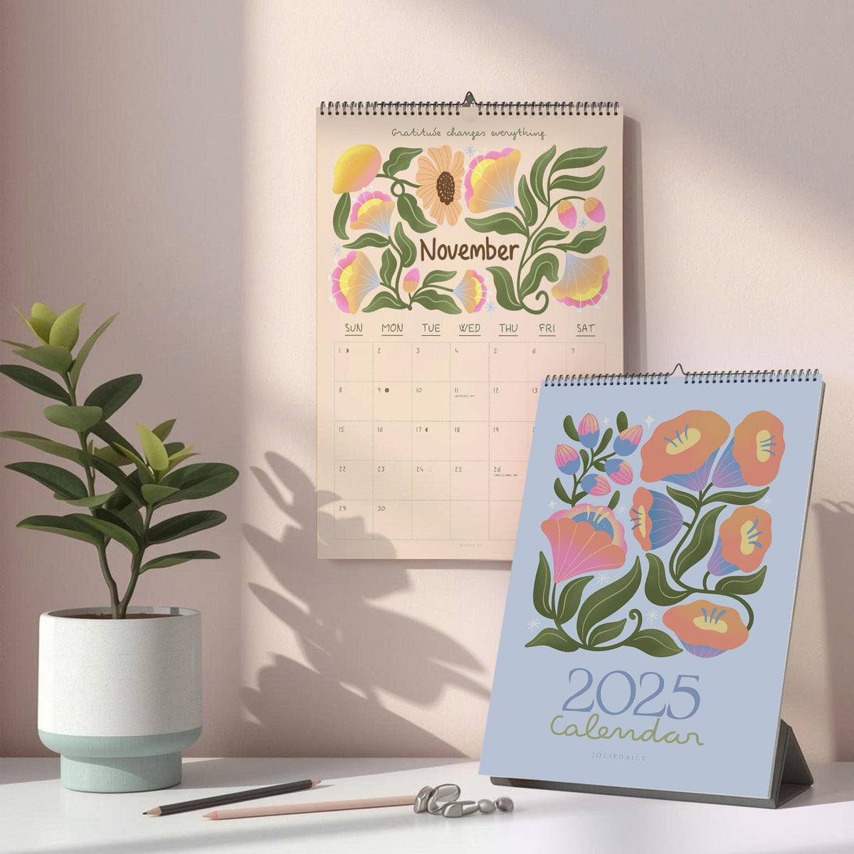 May include: Two calendars, one hanging and one standing, with floral designs. The hanging calendar shows the month of November. The standing calendar is a 2025 calendar. A potted plant and pencils are in the foreground.