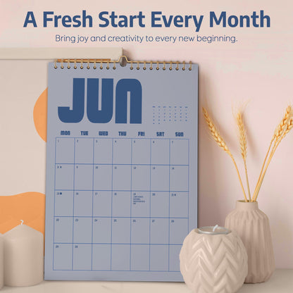 May include: A blue wall calendar for June with gold spiral binding. The calendar has a simple layout with the days of the week across the top. The text "A Fresh Start Every Month" is at the top. A vase with wheat stalks and a candle holder are in the foreground.