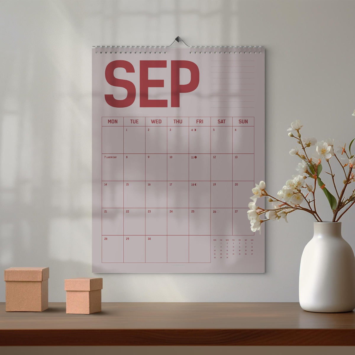 May include: A wall calendar for September with a red and white design. The calendar is hanging on a white wall. Two small, brown cardboard boxes sit on a wooden table in front of the calendar. A white vase with white flowers is to the right.