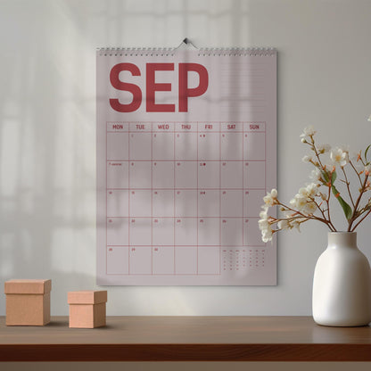 May include: A wall calendar for September with a red and white design. The calendar is hanging on a white wall. Two small, brown cardboard boxes sit on a wooden table in front of the calendar. A white vase with white flowers is to the right.