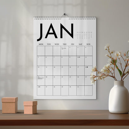 May include: A white wall calendar for January, with the month's name in large black letters. The calendar grid is simple, with days of the week across the top. Two small, brown cardboard boxes sit on a wooden surface below the calendar.