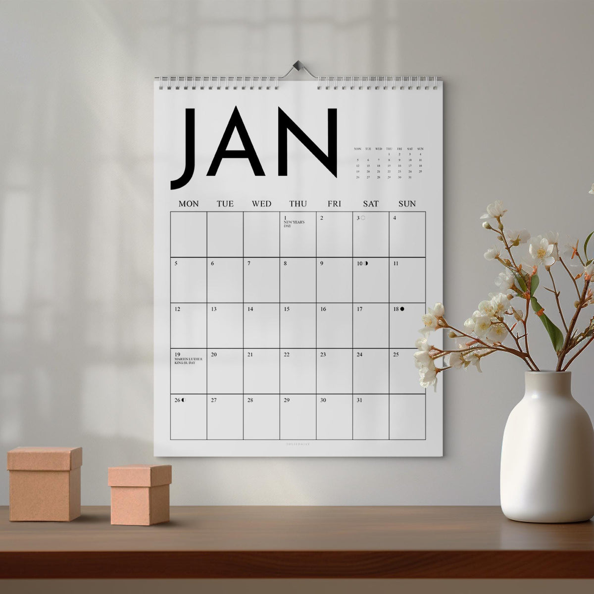 May include: A white wall calendar for January, with the month's name in bold black letters. The calendar grid is simple, with days of the week across the top. Two small, brown cardboard boxes sit on a wooden surface below the calendar.