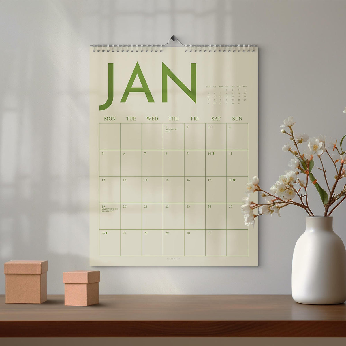 May include: A wall calendar for January with green lettering and a spiral binding. The calendar is hanging on a white wall above a wooden table. Two small, brown cardboard boxes sit on the table, and a white vase with flowers is to the right.