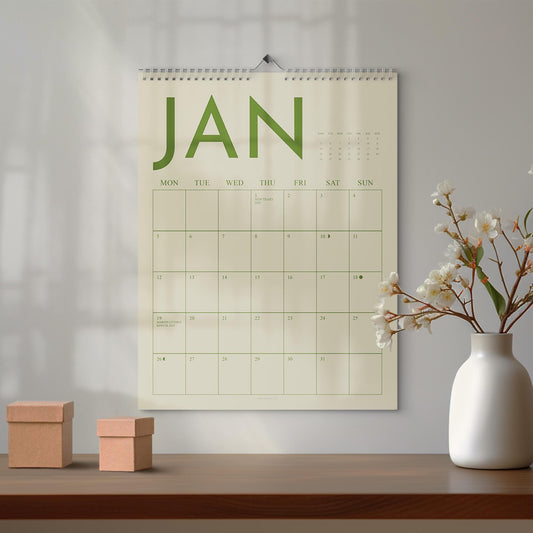 May include: A wall calendar for January with green lettering and a spiral binding. The calendar is hanging on a white wall above a wooden table. Two small, brown cardboard boxes sit on the table, and a white vase with flowers is to the right.