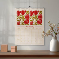 May include: A wall calendar for February with a floral design in red and olive green. The calendar is hanging on a wall above a wooden table. Two small, square cardboard boxes sit on the table, and a white vase with flowers is to the right.