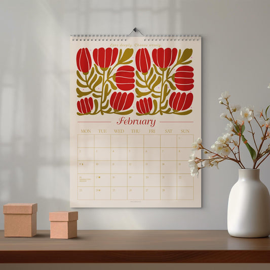 May include: A wall calendar for February with a floral design in red and olive green. The calendar is hanging on a wall above a wooden table. Two small, square cardboard boxes sit on the table, and a white vase with flowers is to the right.